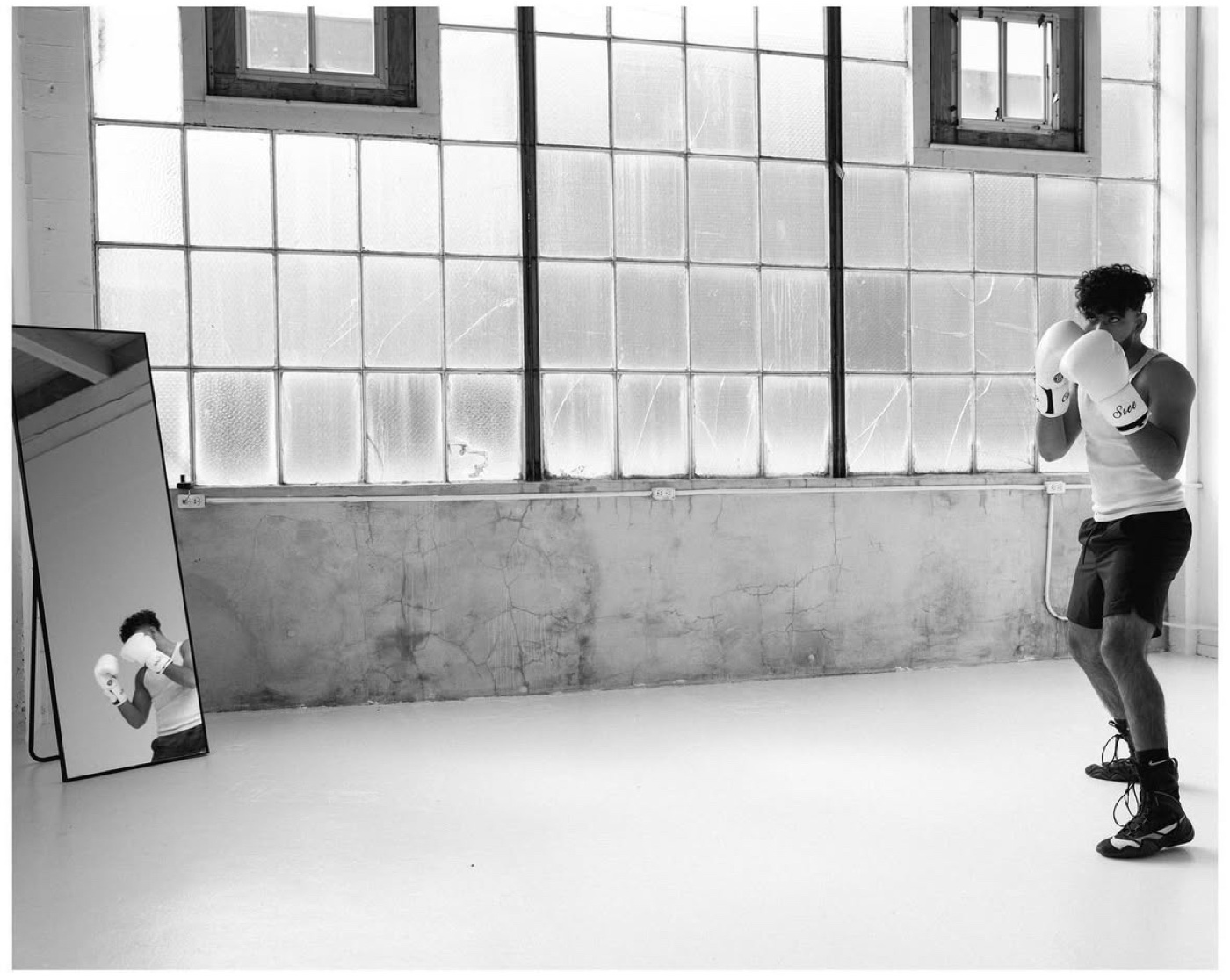 Sree shadowboxing beside tall factory windows, reflected in a floor mirror