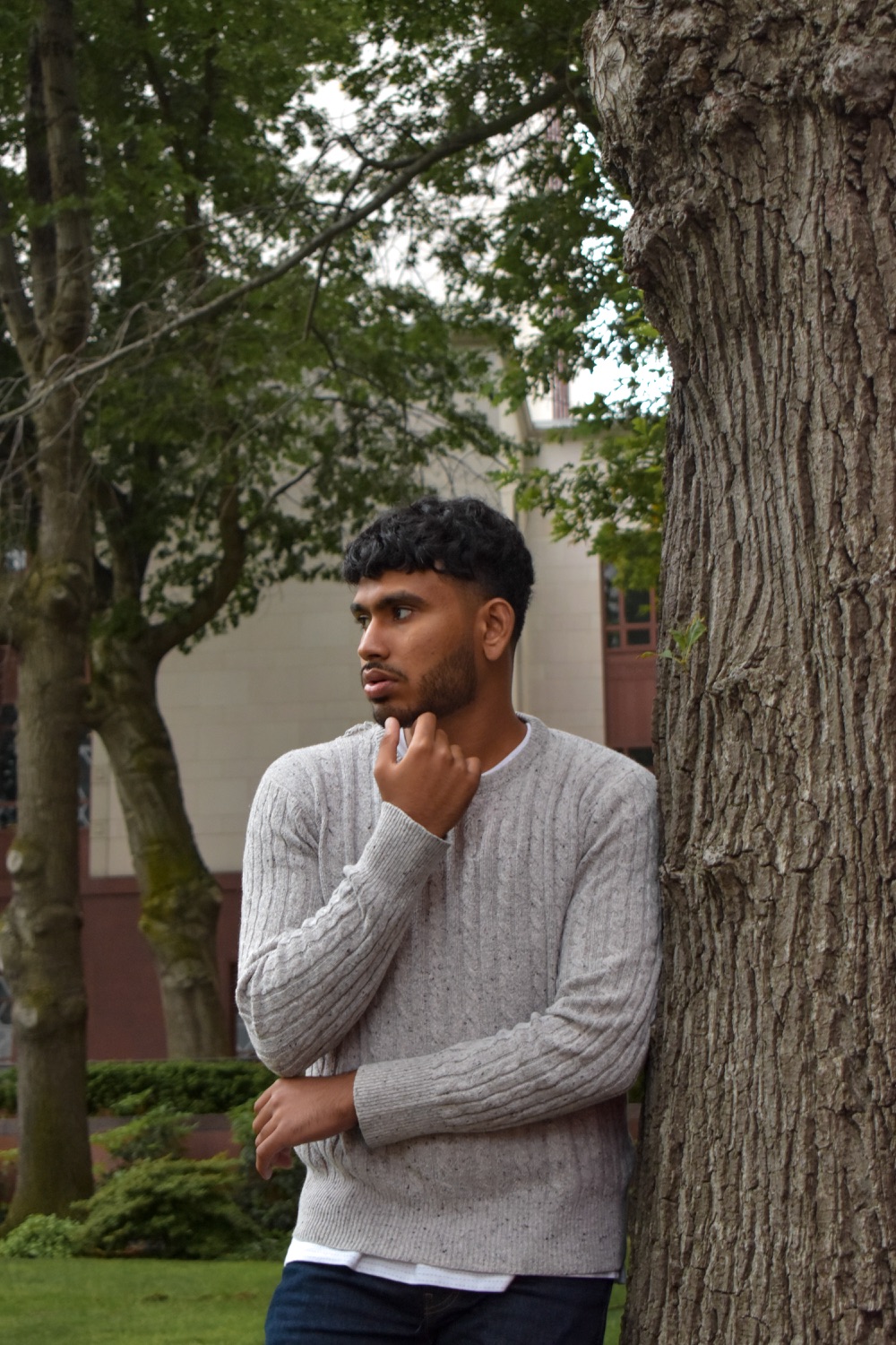 Sree in a grey cable-knit sweater leaning against a tree, hand at his chin